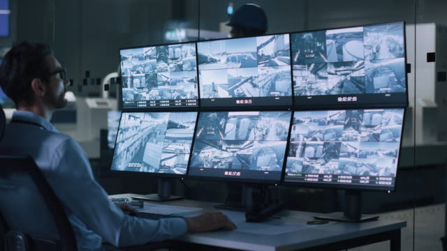 Industry 4.0 Modern Factory: Security Operator Controls Proper Functioning of Workshop Production Line, Uses Computer with Screens Showing Surveillance Camera Footage Feed. High-Tech Security