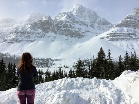 A young woman admiring an incredible view of a dark green forest in the foreground and snow covered mountains peaks in the background, along the icefield parkway in the Rocky Mountains Alberta, Canada