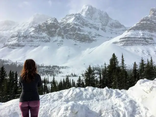 Photo of A young woman admiring an incredible view of a dark green forest in the foreground and snow covered mountains peaks in the background, along the icefield parkway in the Rocky Mountains Alberta, Canada