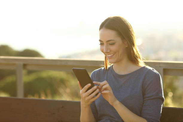 istock Happy woman using mobile phone sitting on a bench 1208292952