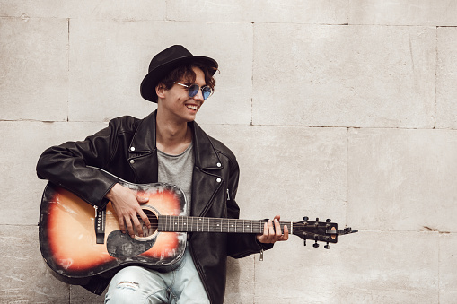 Young guy musician wearing hat and sunglasses standing leaning back on wall playing guitar looking aside laughing cheerful