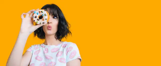 Charming brunette girl looking through a donut is feeling amazed posing on a yellow background with freespace