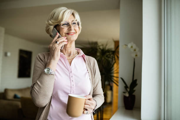 femme aînée heureuse parlant au téléphone tout en buvant le café par la fenêtre. - women telephone senior adult on the phone photos et images de collection