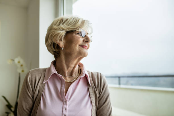 istock Smiling senior woman day dreaming while looking through the window. 1208325021