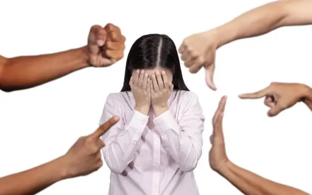 Photo of woman covered her face with regret for being bullying , sexism , racism and hate surrounded by hands mocking her, scoffing with offended at social of workplace