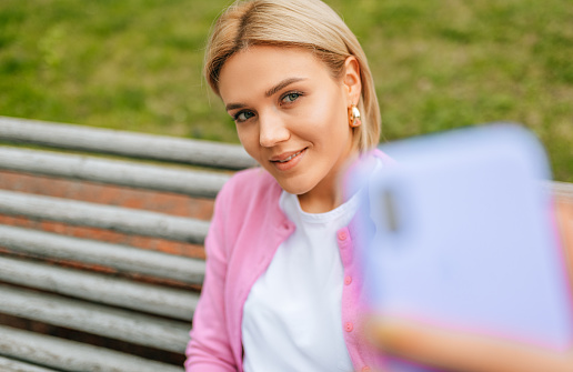 Gorgeous blonde young woman wearing blue jeans, white t-shirt and pink jacket, smiling and taking selfie on smart phone. Happy female making self portrait in the city street in spring time.