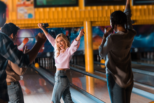 excited blonde girl showing winner gesture while standing near bowling alley with multicultural friends