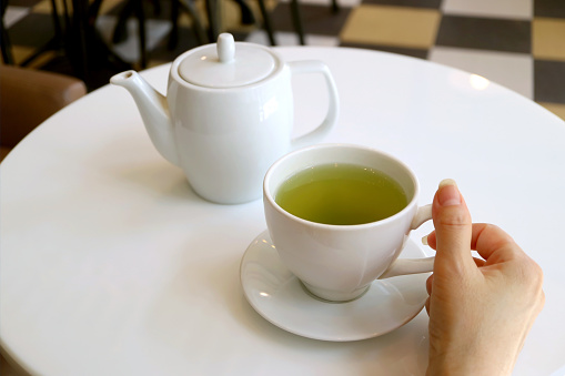 Female's hand holding cup of green tea on a white round table with teapot in background