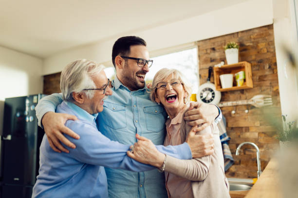 Cheerful senior couple and their adult son having fun while embracing at home. Cheerful mid adult man and his senior parents laughing while embracing in the kitchen. reunion stock pictures, royalty-free photos & images