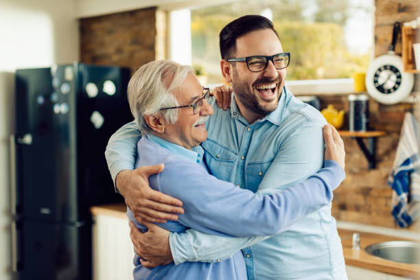 Cheerful mid adult man and his senior father embracing in the kitchen. Cheerful mature man and his adult son embracing while greeting in the kitchen. son stock pictures, royalty-free photos & images