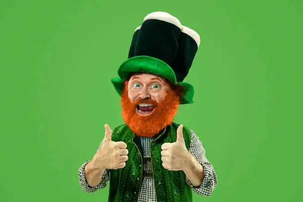 Nice, thumbs up. Excited leprechaun in green suit with red beard on green background. Funny portrait of man ready to party. Saint Patrick day, human emotions, celebration, traditional holidays.