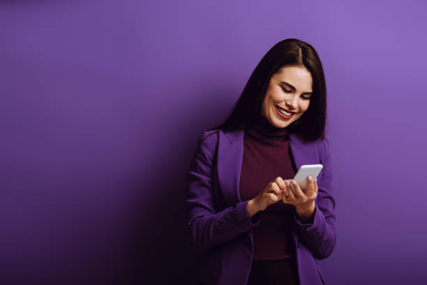 istock cheerful young woman smiling while chatting on smartphone on purple background 1208340393