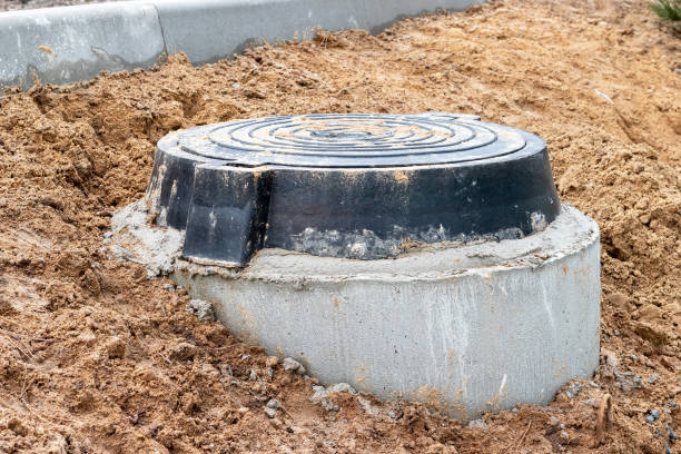 A black cast-iron sewer hatch on a concrete base, located on a mound of damp ground for access to the sewer well at the construction site of a new residential complex