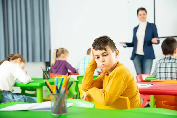 Portrait of upset boy in schoolroom on background with pupils studying with teacher