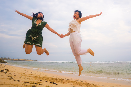 young beautiful and happy couple of attractive Asian Korean women jumping on the air together at the beach enjoying holidays having crazy fun feeling free and joyful  in girls friendship concept