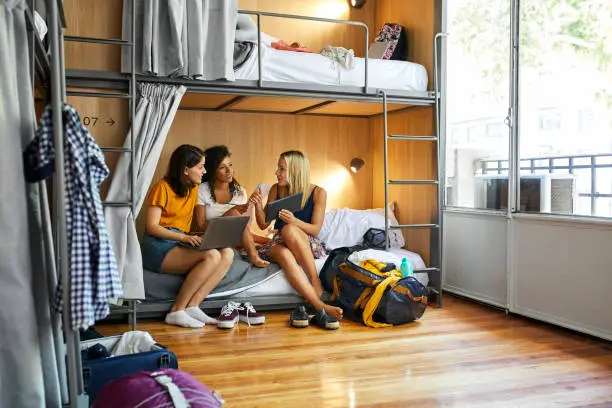 Female friends discussing while sitting on bunkbed. Young women are using technologies and talking over map. They are planning trip at dorm room in hostel.