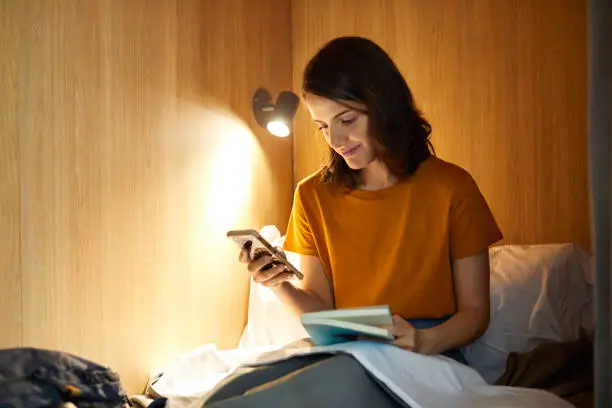 Smiling young woman using mobile phone while reading book. Female is sitting on illuminated bunkbed. She is in casuals at hostel during vacation.