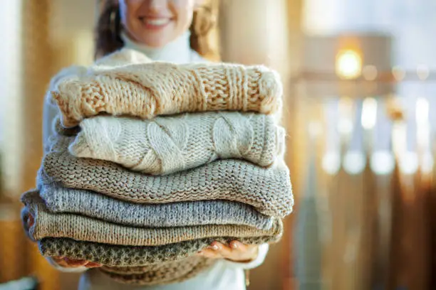 Closeup on happy elegant middle age housewife in white sweater and skirt at modern home in sunny winter day holding pile of sweaters.