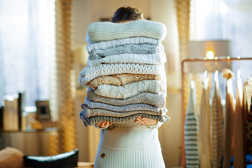 modern woman in white sweater and skirt in the modern house in sunny winter day holding huge pile of sweaters.