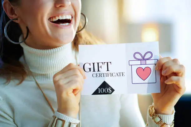 Closeup on smiling modern 40 years old woman in white sweater and skirt at modern home in sunny winter day holding gift certificate.
