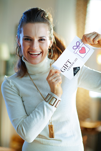 Portrait of happy young woman in white sweater and skirt in the modern house in sunny winter day showing gift certificate.
