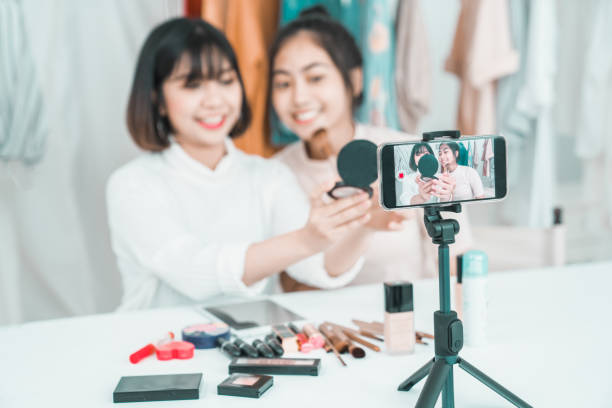 Two young girls doing video recording by smartphone camera. Asian woman make up artist applying powder on female blogger face with cosmetic products table in living room. Beauty ladies live stream Two young girls doing video recording by smartphone camera. Asian woman make up artist applying powder on female blogger face with cosmetic products table in living room. Beauty ladies live stream tutorial photos stock pictures, royalty-free photos & images