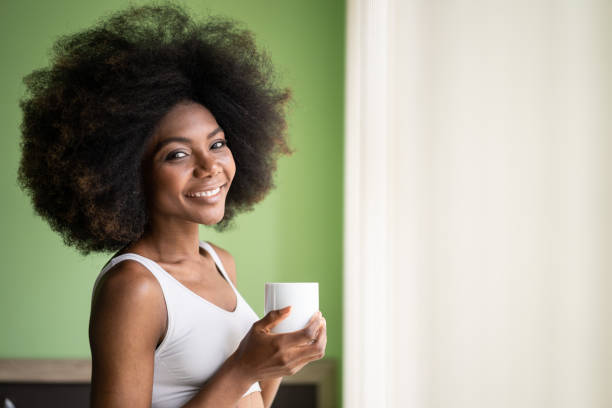 istock Smiling afro woman looking through the window with a cup of coffee 1208367351