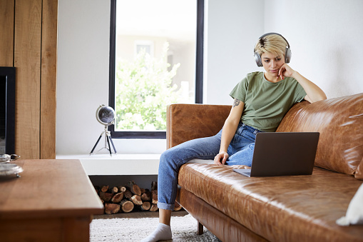 Young A woman watching movie in living room. Female is wearing headphones while using laptop. She is sitting on sofa at home.