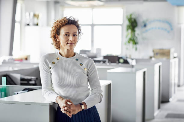 Confident businesswoman with hands clasped Portrait of confident mature female entrepreneur at workplace. Blond Professional is leaning on cabinet at office. Businesswoman is standing with hands clasped. middle aged woman fashion stock pictures, royalty-free photos & images