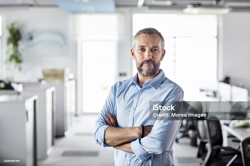 Confident male professional with arms crossed Confident male professional with arms crossed at workplace. Portrait of bearded mature businessman. Entrepreneur is standing at office. Men Stock Photo