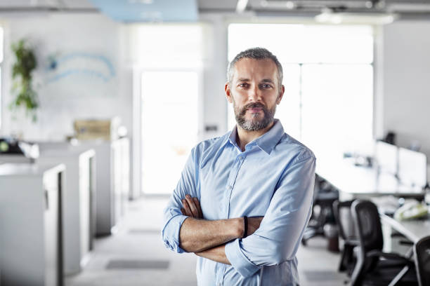 Confident male professional with arms crossed Confident male professional with arms crossed at workplace. Portrait of bearded mature businessman. Entrepreneur is standing at office. 40 44 years stock pictures, royalty-free photos & images