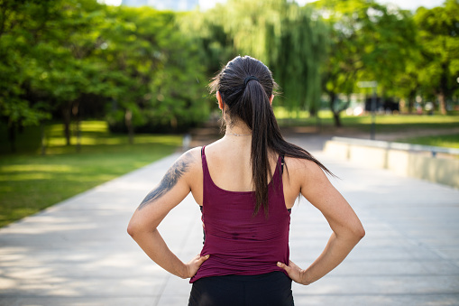 Rear view of tattooed Latin woman in sport clothes standing in park, preparing for jogging session on beautiful sunny morning