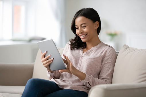 Smiling young African American woman sit relax on couch in living room browsing Internet shopping online on tablet, happy biracial female rest on sofa at home texting messaging on modern pad gadget