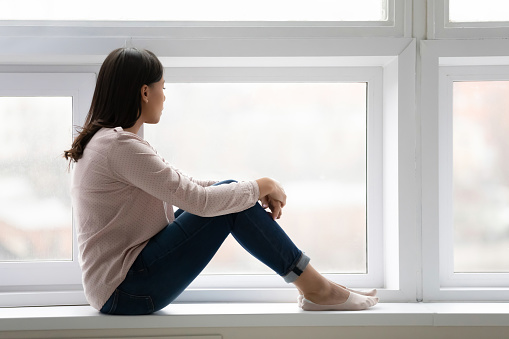 Thoughtful African American young woman sit on windowsill at home look in distance pondering or thinking, pensive unhappy biracial female lost in thoughts feel lonely, dreaming or visualizing