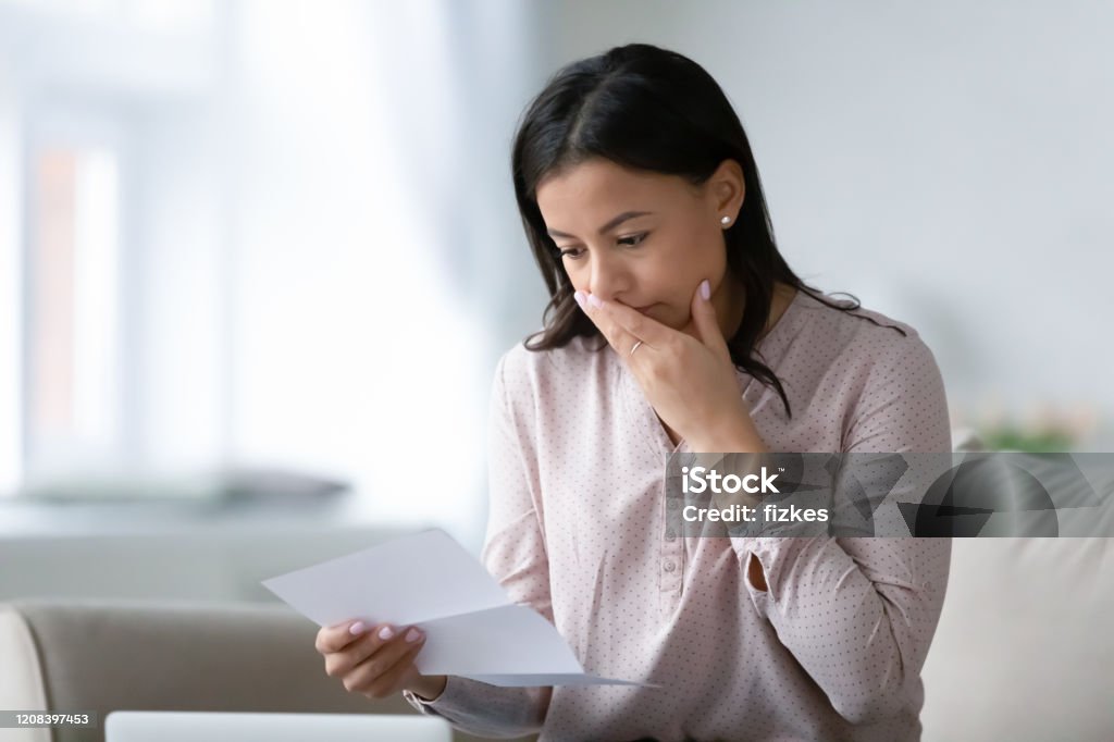 Unhappy biracial woman read bad news in letter Distressed African American young woman sit on couch at home read bad negative news in paper correspondence, unhappy biracial female stressed consider unpleasant message in postal letter Financial Bill Stock Photo