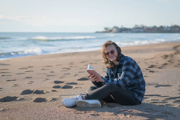 istock Handsome man sitting on a beach and reading a book 1208401951