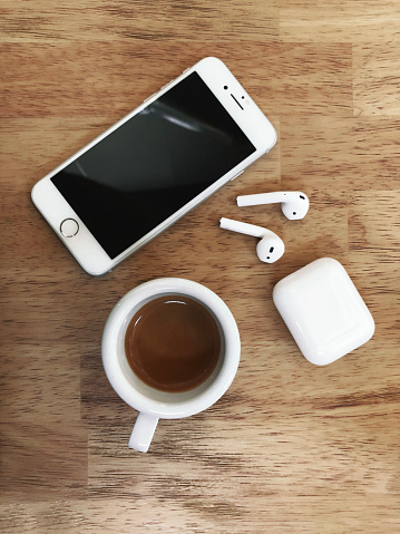 Izmir, Turkey - July  11, 2019: Top view of White colored Apple brand Iphone 8 Phone and Airpods 2 on a wooden table with charging box and cup of espresso on a wooden table.