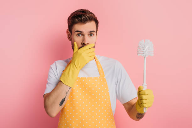 shocked young man in apron and rubber gloves covering mouth with hand while holding toilet brushon pink background shocked young man in apron and rubber gloves covering mouth with hand while holding toilet brush on pink background toilet brush stock pictures, royalty-free photos & images