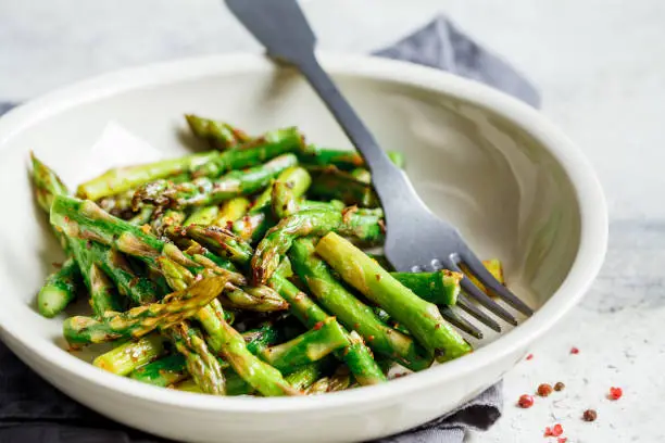 Photo of Cooked green asparagus with pepper and salt in a white bowl.