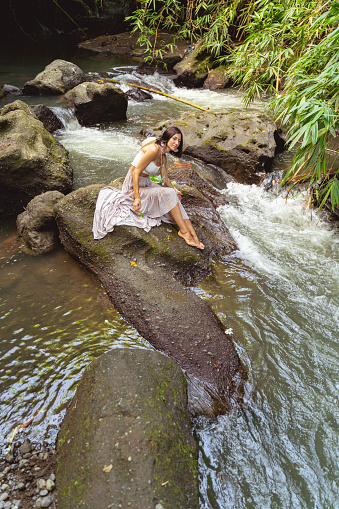 Joyful young female is sitting on stone in mountain river and enjoying connection with exotic nature