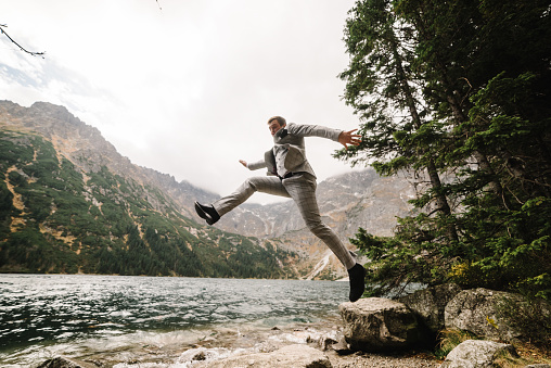 Man jump on the stony shore. The groom near the lake in the mountains. Groom against the backdrop of a mountain landscape.