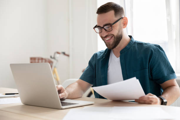 feliz joven hombre de negocios en anteojos trabajando en la computadora. - looking at papers fotografías e imágenes de stock