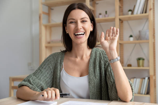 Happy young 30s woman waving hello to camera. Head shot happy young 30s woman sitting at wooden desk, looking at camera, waving hello. Excited businesswoman teacher lecturer recording educational video, greeting students at online workshop. waving gesture stock pictures, royalty-free photos & images