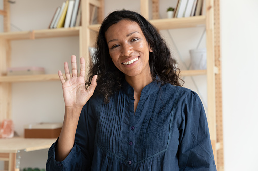 Head shot close up pleasant smiling biracial woman making hello gesture, looking at camera. Positive young african american teacher tutor coach greeting clients online before educational webinar.