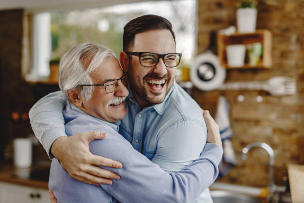 Cheerful man and his senior father embracing while greeting in the kitchen. Happy mature man having fun while embracing with his adult son who came to visit him. offspring stock pictures, royalty-free photos & images