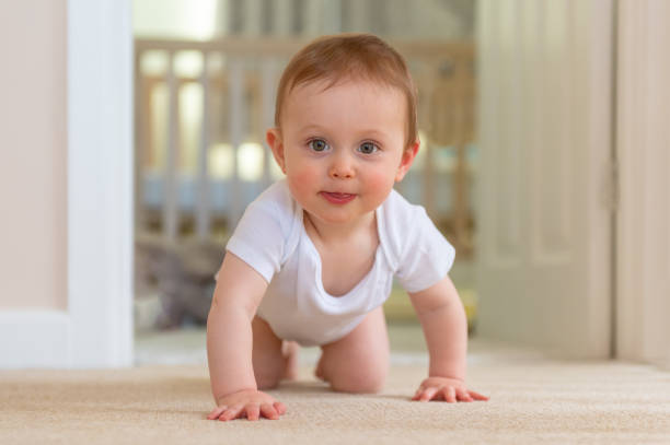 Baby boy in white sleep vest crawling towards the camera on carpet floor.