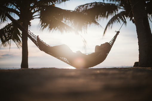 A young man is lying in a hammock at sunset by the ocean. Handsome guy is resting in a hammock among palm trees on the seashore.