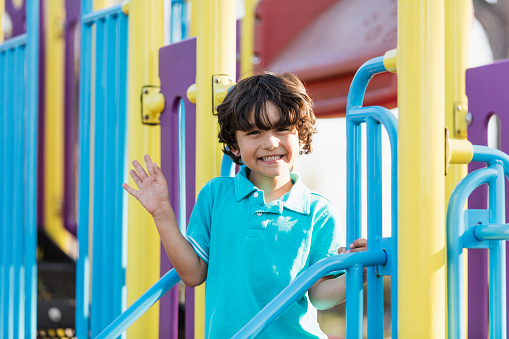 A 6 year old Hispanic boy having fun at the park, standing on playground equipment, smiling and waving at the camera on a sunny spring or summer day.
