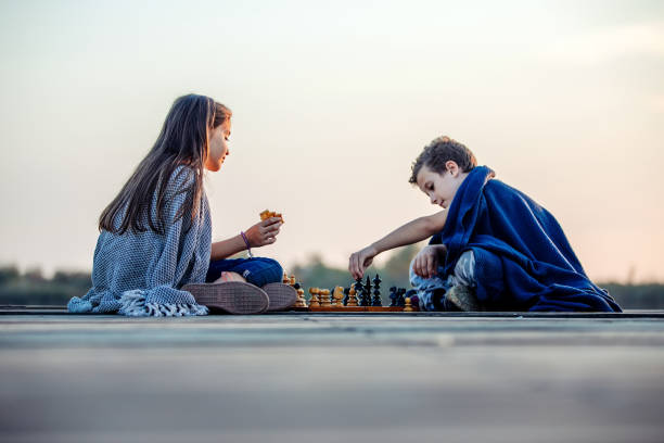 deux jeunes petits amis mignons, garçon et fille ayant l’amusement tout en jouant aux échecs s’asseyant au bord du lac dans la soirée. - brain case photos et images de collection