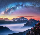 Milky Way above mountains in fog at night in summer. Landscape with alpine mountain valley, low clouds, colorful starry sky with milky way at sunset. Aerial view. Passo Giau, Dolomites, Italy. Space